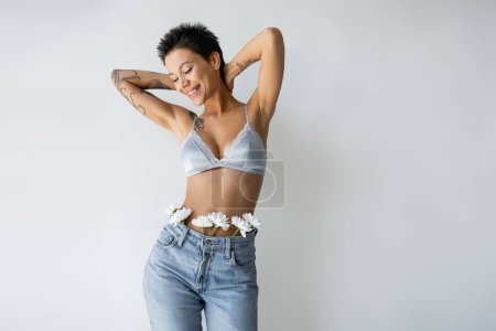 joyful and sexy woman in satin bra and blue jeans with white flowers standing with hands behind neck isolated on grey