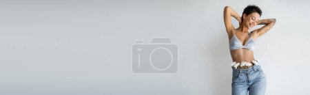 Téléchargez les photos : Heureuse femme tatouée portant un soutien-gorge bleu et un jean avec des fleurs blanches tenant les mains derrière le cou isolé sur gris, bannière - en image libre de droit