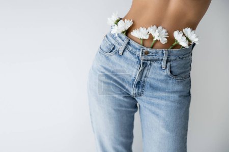 Téléchargez les photos : Partial view of shirtless woman with white flowers in blue jeans isolated on grey - en image libre de droit