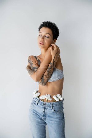 sensual tattooed woman in bra and jeans with white flowers posing with crossed arms and closed eyes isolated on grey