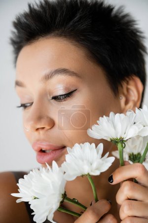 Téléchargez les photos : Portrait en gros plan de jeune femme brune avec maquillage tenant des fleurs blanches isolées sur gris - en image libre de droit
