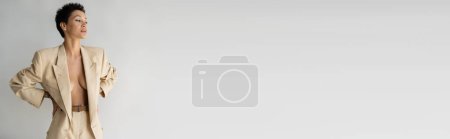Téléchargez les photos : Sexy femme torse nu en blazer beige debout avec les mains sur la taille et regardant loin isolé sur gris, bannière - en image libre de droit