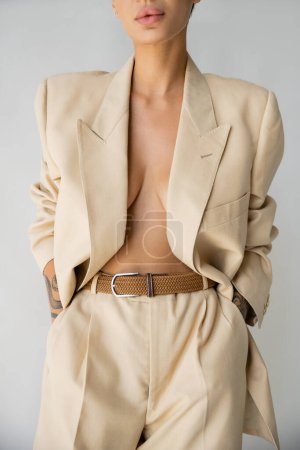 cropped view of sexy woman wearing blazer on shirtless body and holding hands in pockets of trousers isolated on grey