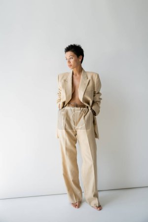 full length of sexy barefoot woman in beige oversize blazer standing with hands in pockets and looking away on grey background