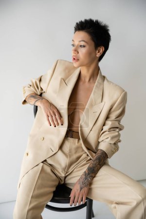sensual tattooed woman in stylish blazer on shirtless body sitting on chair and looking away on grey background