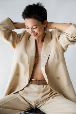 smiling woman in oversize blazer on shirtless body posing with hands behind neck isolated on grey