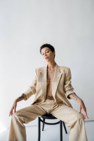 Photo for Sexy shirtless woman in trendy oversize suit sitting on chair and looking at camera on grey background - Royalty Free Image