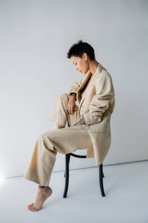 Téléchargez les photos : Side view of barefoot brunette woman in suit sitting on chair on grey background - en image libre de droit