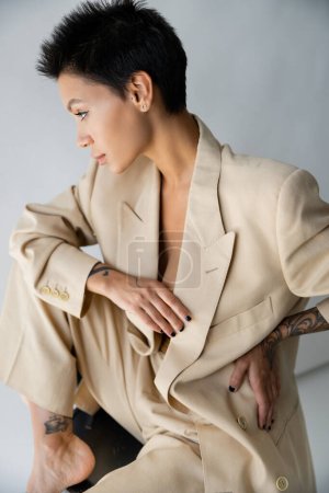 stylische brünette Frau hält die Hand auf der Hüfte, während sie auf einem Stuhl auf grauem Hintergrund sitzt