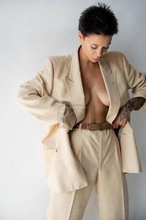 young woman in beige blazer on shirtless body adjusting oversize pants on grey background