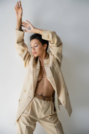 jeune et sexy femme en costume beige sur corps torse nu posant avec les bras vers le haut sur fond gris