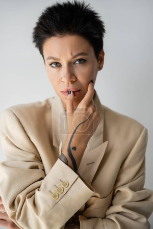 Téléchargez les photos : Portrait of thoughtful tattooed woman in beige blazer touching face and looking at camera isolated on grey - en image libre de droit