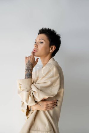 Photo for Thoughtful tattooed woman in beige blazer holding hand near chin and looking away on grey background - Royalty Free Image