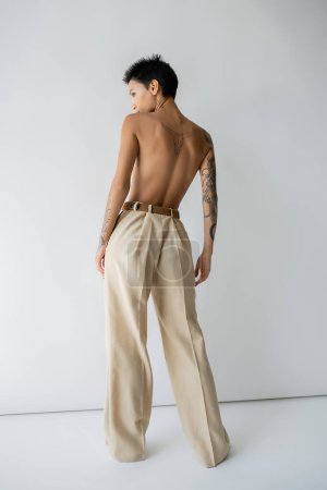 back view of shirtless tattooed woman in oversize pants standing and looking away on grey background