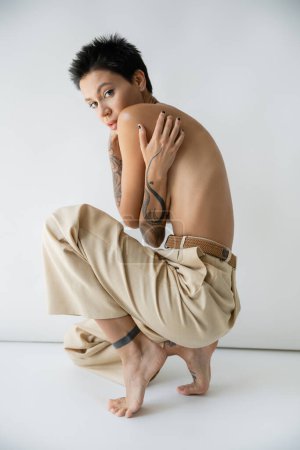 femme séduisante torse nu dans un pantalon élégant regardant la caméra tout en s'étreignant sur fond gris