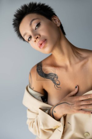 portrait of sexy tattooed woman with naked shoulders covering breast and looking at camera isolated on grey