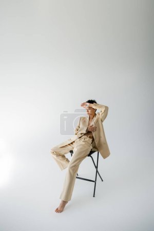 full length of barefoot woman in beige suit sitting on chair and holding hand near forehead on grey background