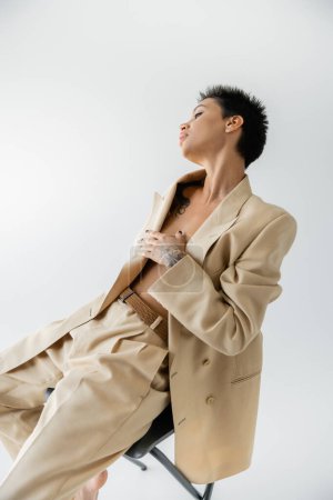 sexy hemdlose Frau, die die Brust mit der Hand bedeckt, während sie im beigefarbenen Oversize-Anzug isoliert auf grau sitzt