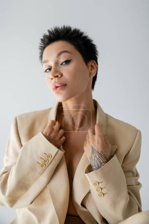 portrait of pretty woman with piercing and makeup posing in beige blazer isolated on grey