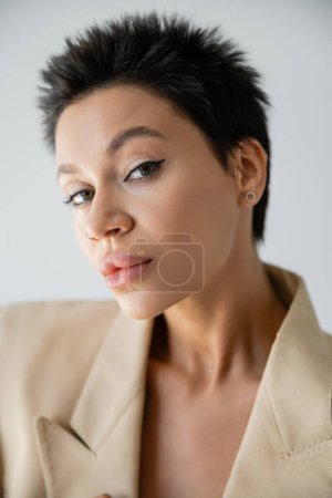 Téléchargez les photos : Portrait de femme branchée aux cheveux bruns courts et maquillage regardant la caméra isolée sur gris - en image libre de droit