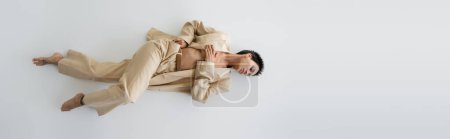 top view of barefoot and shirtless woman in beige pantsuit lying on grey background, banner