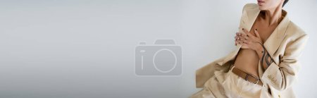 Photo for Partial view of tattooed woman covering bust with hands while sitting in pantsuit on grey background, banner - Royalty Free Image