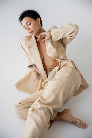 Foto de Young and sexy woman in beige suit on shirtless body sitting on grey background - Imagen libre de derechos