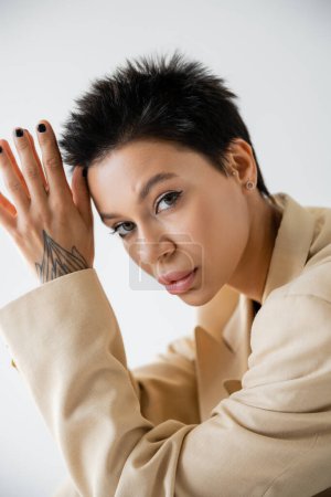 Foto de Retrato de mujer morena tatuada con maquillaje posando con las manos cerca de la cabeza y mirando a la cámara aislada en gris - Imagen libre de derechos