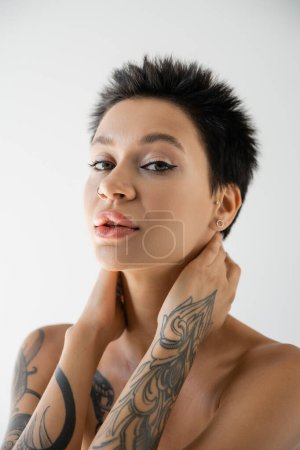 portrait of tattooed woman with makeup and bare shoulders holding hands on neck and looking at camera isolated on grey