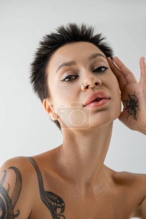portrait of tattooed woman with makeup and piercing holding hand near face isolated on grey