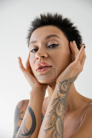 portrait of brunette woman with short hair and makeup holding tattooed hands near face isolated on grey