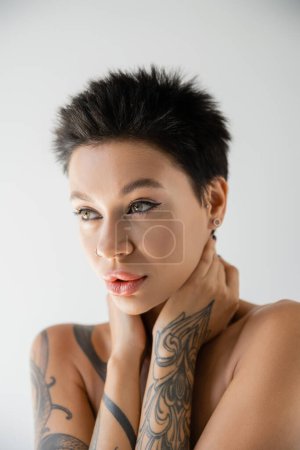 portrait of tattooed brunette woman with makeup touching neck and looking away isolated on grey