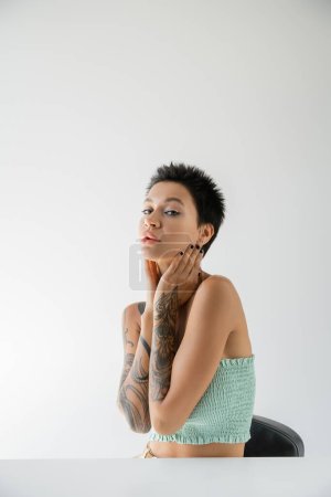 Foto de Young tattooed woman in strapless top holding hands near neck while sitting at table on grey background - Imagen libre de derechos