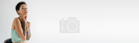 Photo for Tattooed brunette woman in strapless top holding hands near neck and looking at camera isolated on grey, banner - Royalty Free Image