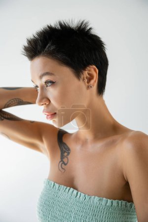 Foto de Joven mujer tatuada en la parte superior sin tirantes sosteniendo la mano cerca de la cabeza y mirando hacia otro lado aislado en gris - Imagen libre de derechos