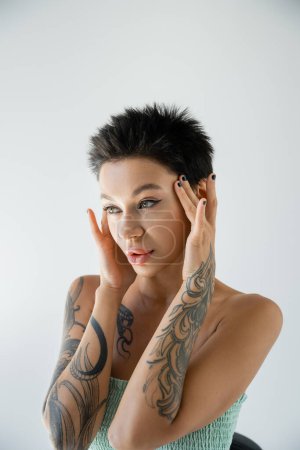 pretty brunette woman with makeup posing with tattooed hands near face and looking away isolated on grey