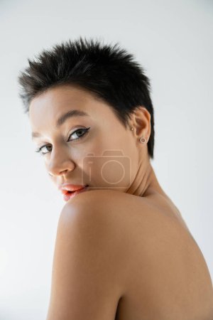 Porträt einer jungen Frau mit kurzen brünetten Haaren und nackter Schulter, die isoliert auf grau in die Kamera blickt
