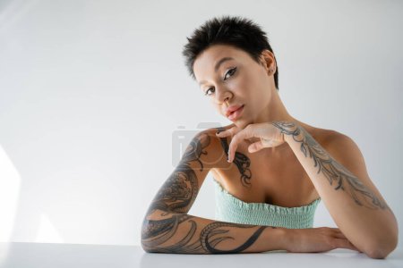 Foto de Sensual mujer tatuada en la parte superior sin tirantes sentado en la mesa y mirando a la cámara sobre fondo gris - Imagen libre de derechos