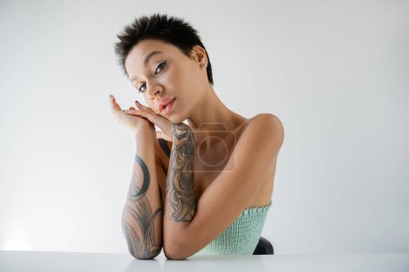 tattooed brunette woman in strapless top sitting with hands near face isolated on grey