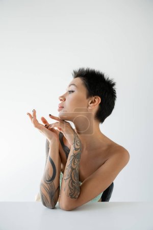 Foto de Retrato de una joven mujer tatuada sentada cerca de la mesa y mirando hacia otro lado aislada en gris - Imagen libre de derechos