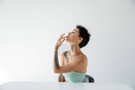 sexy mujer tatuada tocando el labio mientras está sentado cerca de la mesa y mirando hacia otro lado sobre fondo gris
