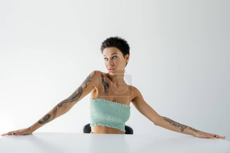Foto de Sexy mujer tatuada en la parte superior sin tirantes sentado en la mesa y mirando hacia otro lado sobre fondo gris - Imagen libre de derechos
