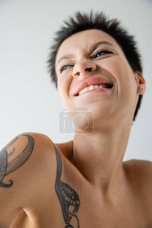 low angle view of excited tattooed woman with piercing biting lip and looking away isolated on grey