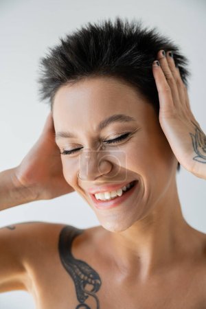 Téléchargez les photos : Portrait d'une femme joyeuse tatouée touchant la tête tout en souriant les yeux fermés isolés sur le gris - en image libre de droit