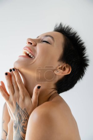excited brunette woman with folded tattooed hands laughing with closed eyes isolated on grey