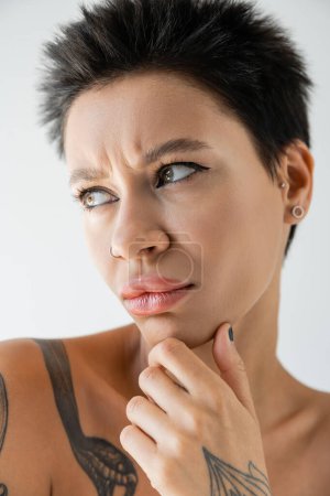 portrait of upset and frowning woman with makeup and piercing touching chin and looking away isolated on grey