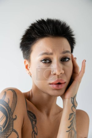 portrait of upset tattooed woman with makeup and piercing touching face and looking at camera isolated on grey