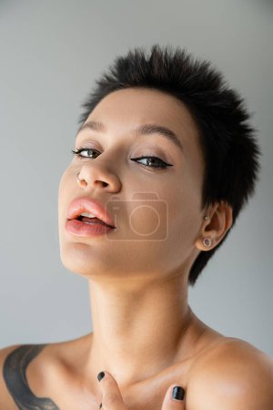 portrait of sexy brunette woman with makeup and piercing looking at camera isolated on grey