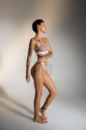 side view of slim brunette woman with tattooed body posing in lingerie on grey background magic mug #638596490