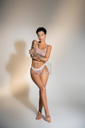 full length of slender tattooed model in underwear standing with crossed arms on grey background